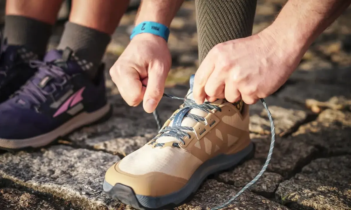 Hände binden Schnürsenkel eines beigefarbenen Schuhs, bereit für den Trail. Daneben ein lila Schuh, der verschiedene wanderschuhe kategorien zeigt.