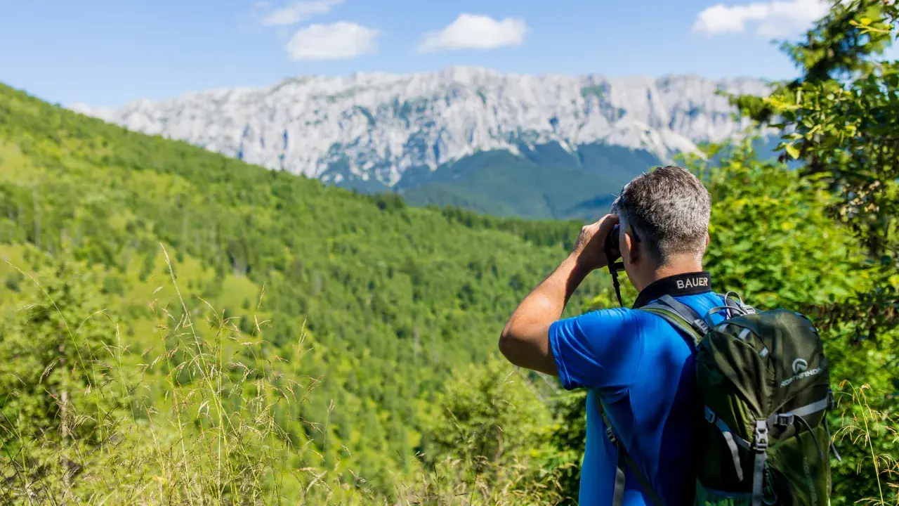 Mann mit Rucksack beim Karpaten Wandern, blickt durch Fernglas auf bewaldete Hänge und felsige Gipfel.