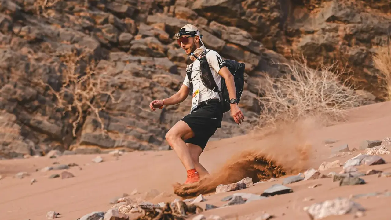 Läufer beim Marathon des Sables wirbelt Sand auf. Ein Mann mit Rucksack und Stirnlampe kämpft sich durch die Wüste.