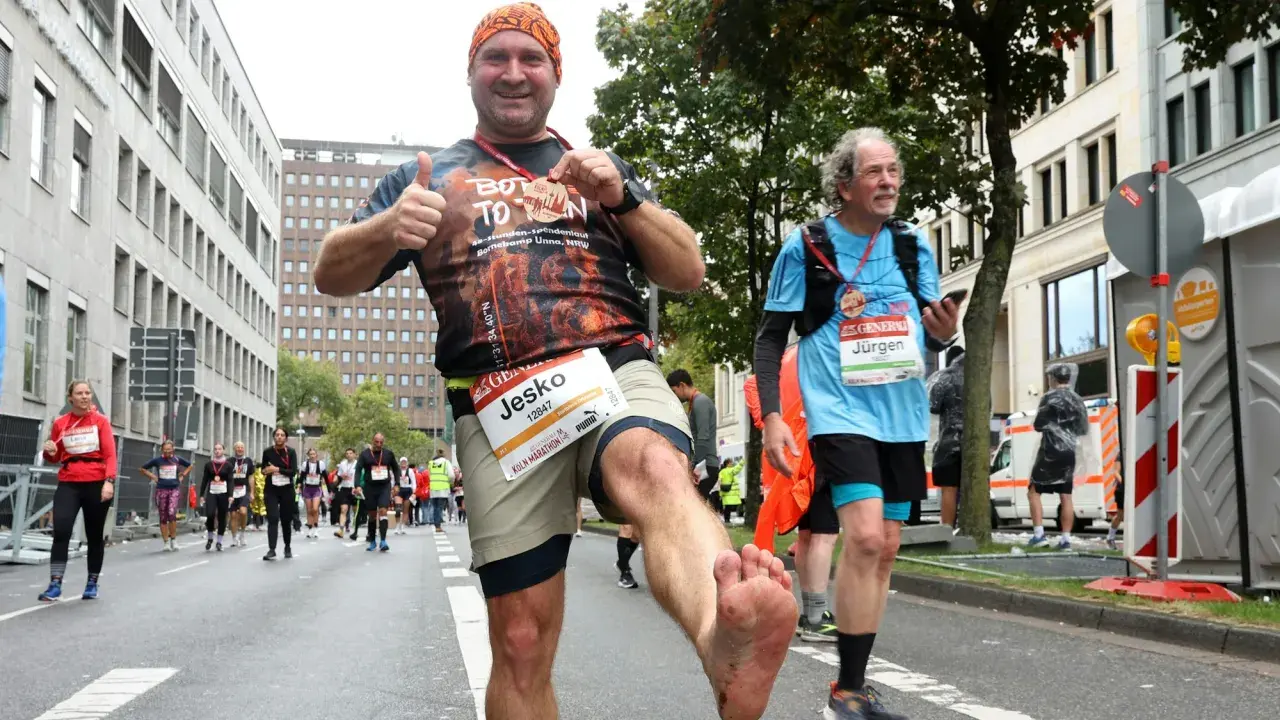 Mann mit schmutzigen Füßen jubelt nach einem Halbmarathon ohne Training. Er hält eine Medaille hoch und gibt Daumen hoch.