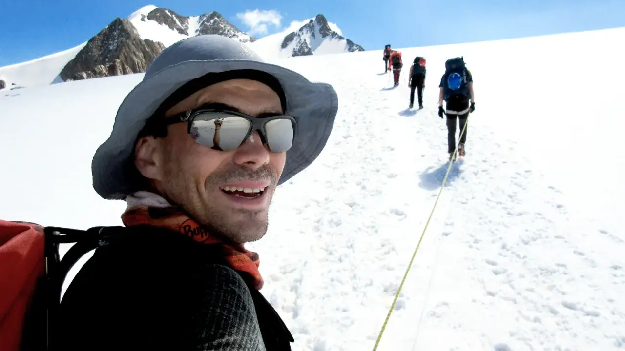 Mann mit Sonnenbrille und Hut, lachend, während er mit einer Gruppe auf einem schneebedeckten Berg wandert. Trail training in den Alpen.