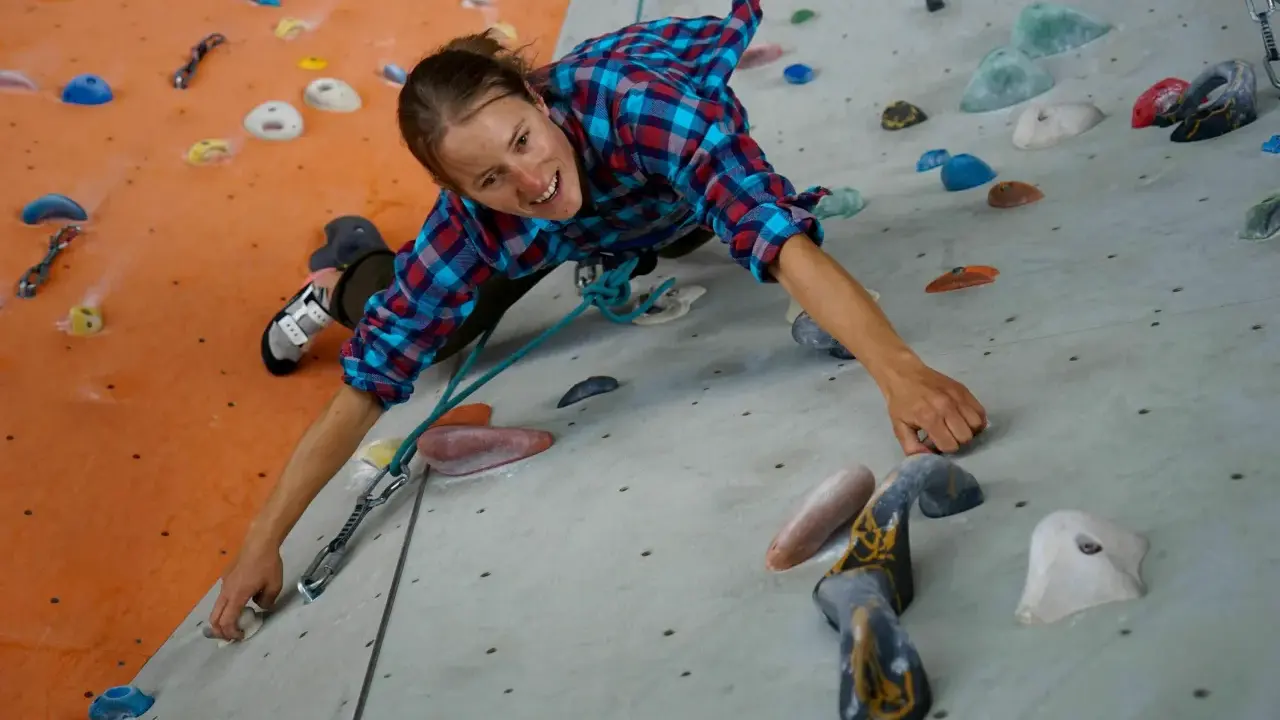 Frau klettert an einer Boulderwand. Sie trägt ein kariertes Hemd und bequeme Kleidung, ideal, was man zum Bouldern anzieht.