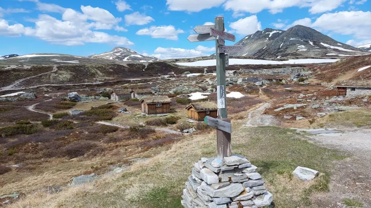 Wanderwegweiser im Rondane Nationalpark, mit grasbewachsenen Hütten und schneebedeckten Bergen im Hintergrund.