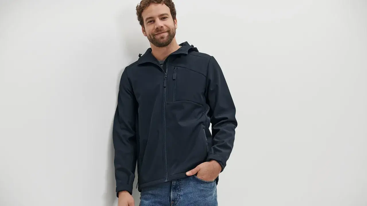 Mann mit lockigem Haar trägt eine dunkelblaue Softshelljacke und Jeans. Die Jacke ist bereit für die Wäsche.