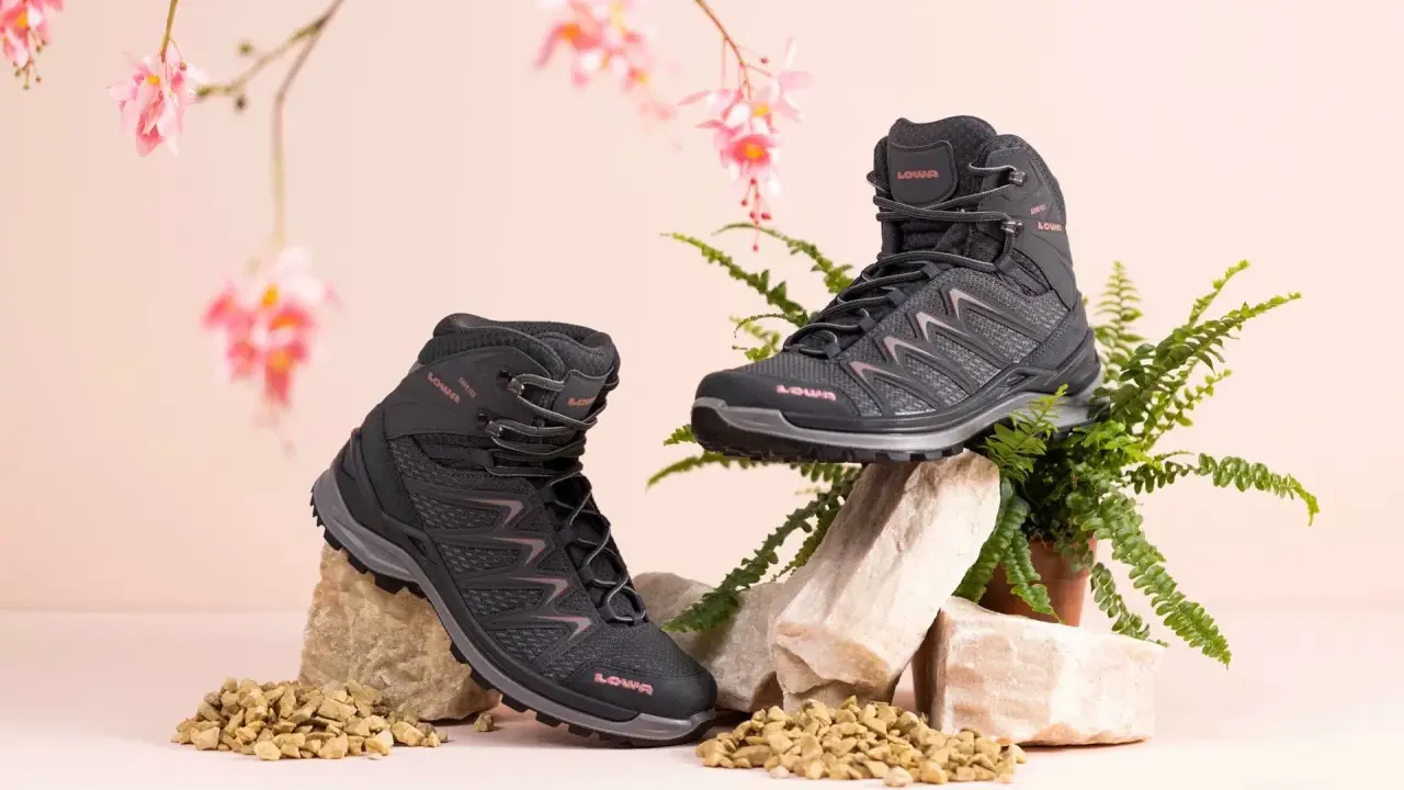 Zwei schwarze Wanderschuhe von LOWA, auf Steinen und Kies arrangiert, mit zarten rosa Blüten und Farn im Hintergrund.