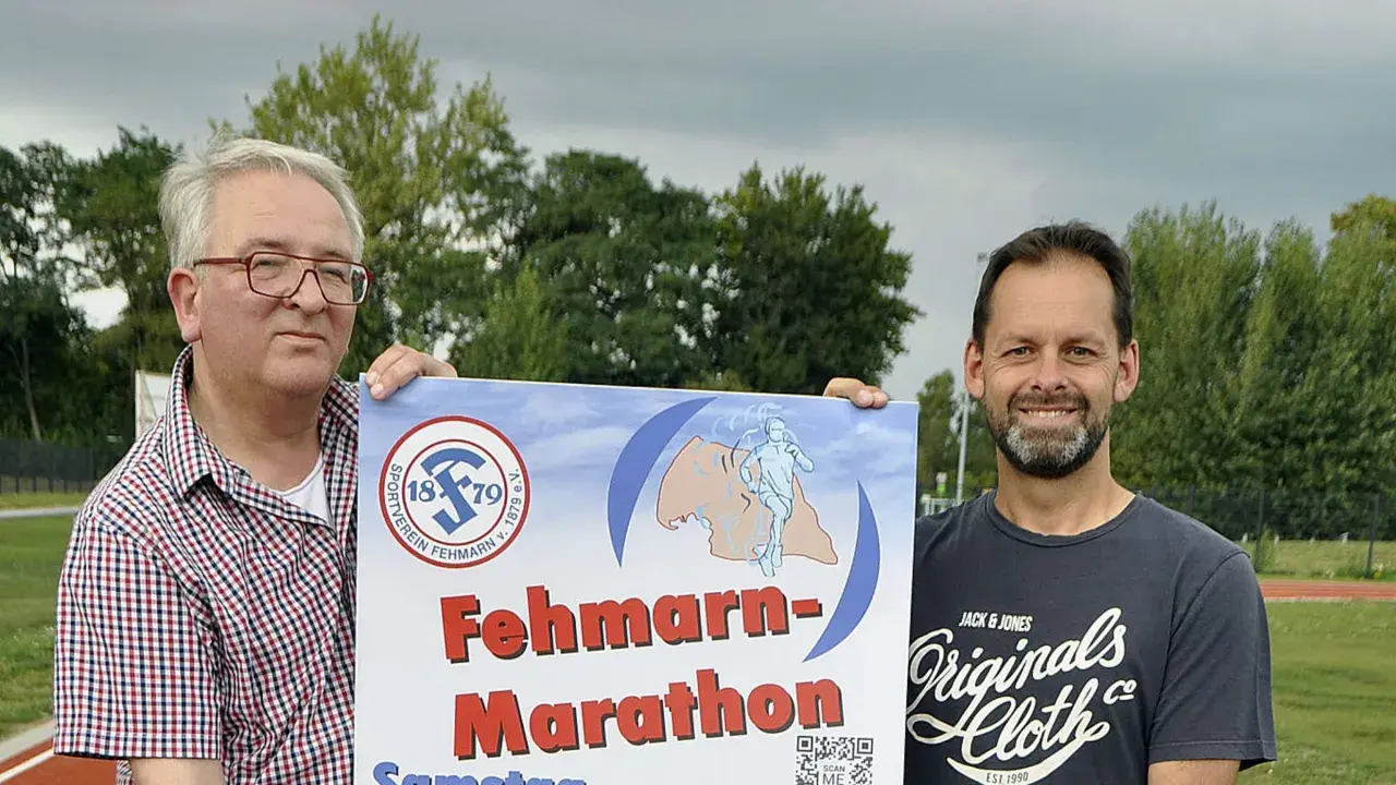 Zwei Männer präsentieren stolz ein Plakat für den Fehmarn Marathon. Das Logo des Sportvereins Fehmarn ist zu sehen.