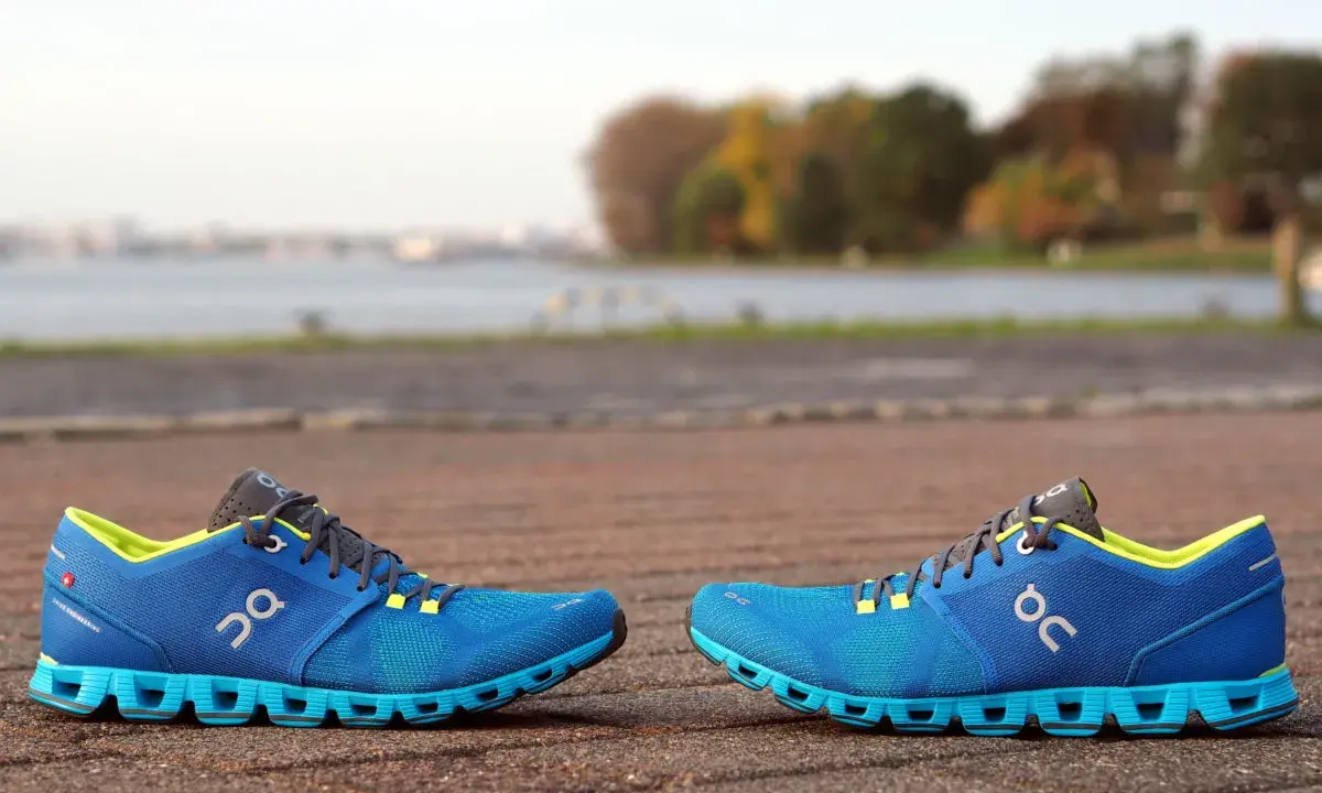 Zwei blaue Laufschuhe, Modell Cloud X4 Damen, stehen auf einem gepflasterten Weg. Im Hintergrund ein See und Bäume.