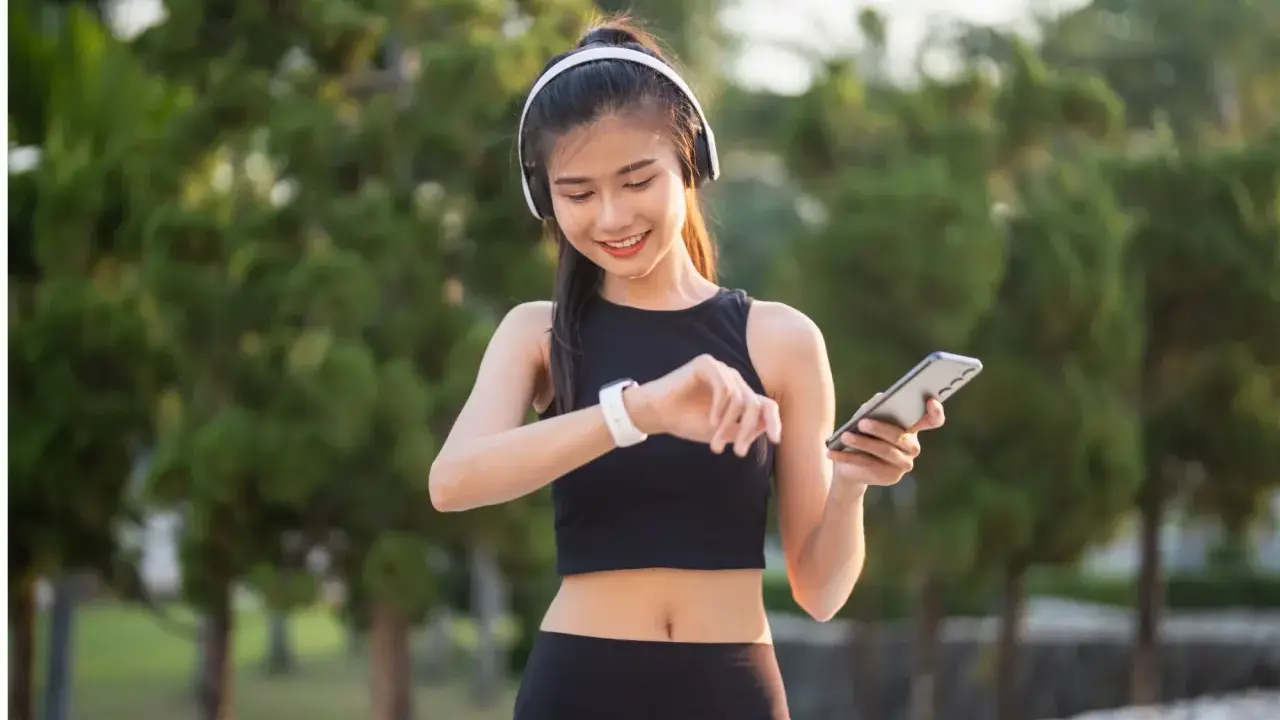 Lächelnde junge Frau mit Kopfhörern und Smartphone, die ihre Smartwatch prüft. Sie trägt atmungsaktive Kleidung für ihr Training im Freien.