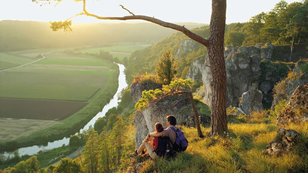 Ein Paar genießt den Sonnenuntergang nach besonderen Wanderungen auf einem Felsvorsprung mit Blick auf ein Tal mit Fluss und Feldern.