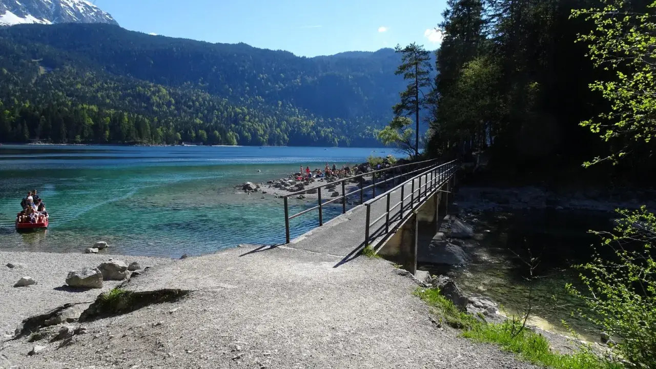 Ein Steg führt über das klare Wasser des Eibsees, ideal für den Eibsee Rundweg. Im Hintergrund Berge und ein rotes Boot.