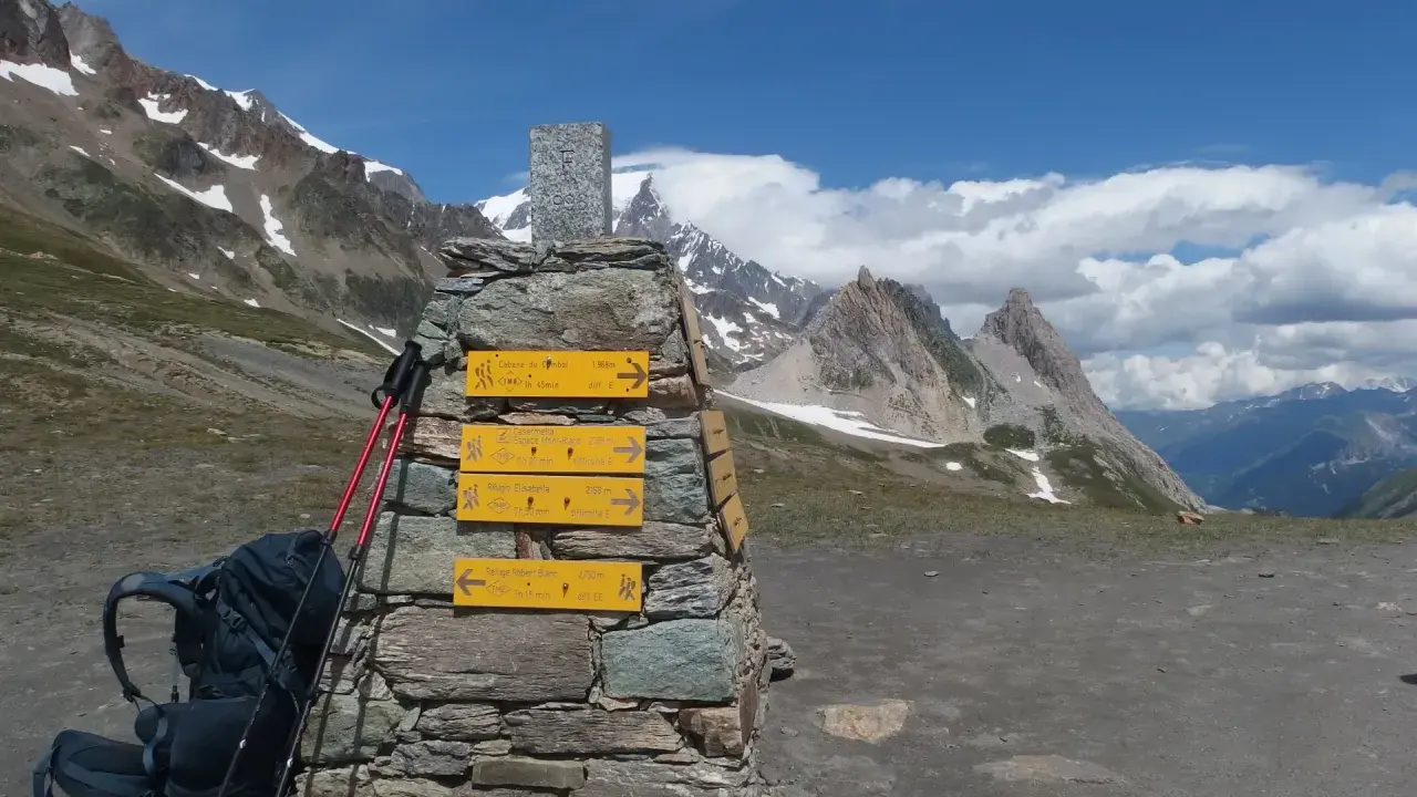 Wegweiser für die Tour du Mont Blanc Etappen: Rucksack und Stöcke liegen bereit. Bergpanorama mit blauen Himmel und Wolken.