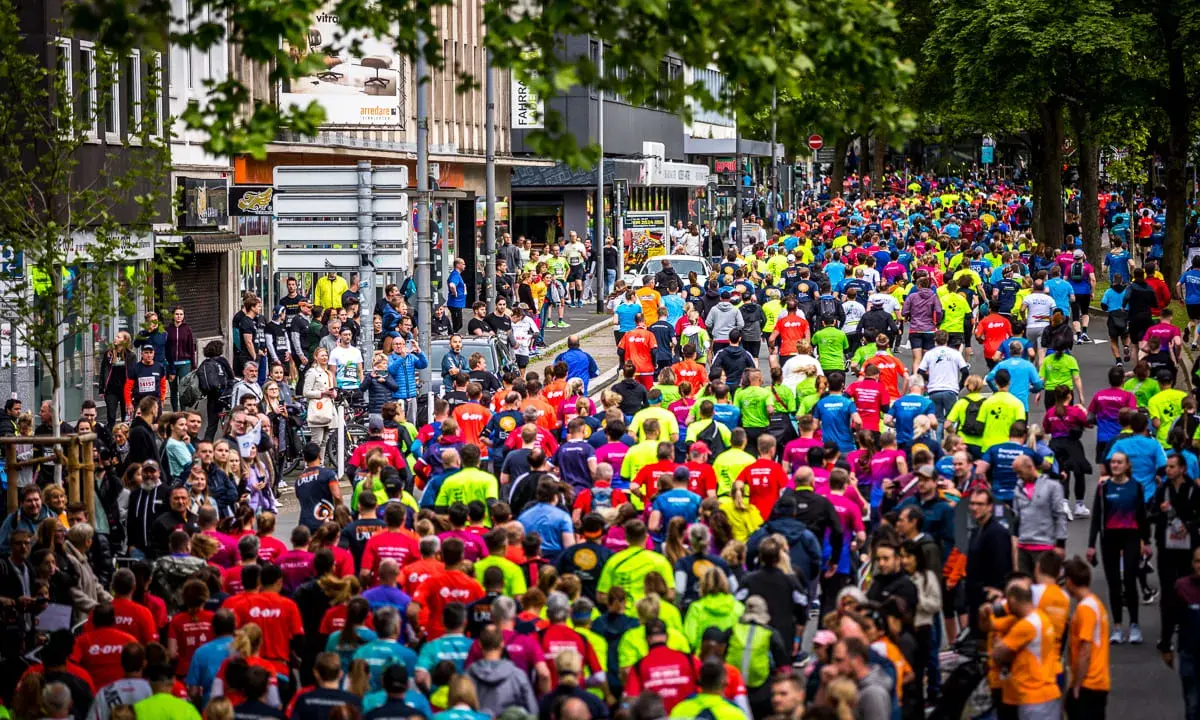 Große Menschenmenge beim Firmenlauf Essen, viele Läufer in bunten Trikots auf einer Straße, umgeben von Bäumen und Gebäuden.