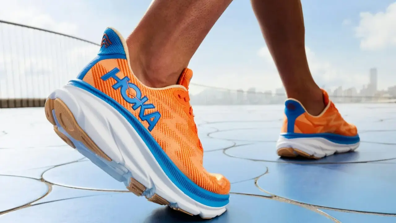 Ein Läufer testet die Hoka Clifton 9 Schuhe in leuchtendem Orange mit blauen Akzenten, die auf einem blauen Boden unterwegs sind.