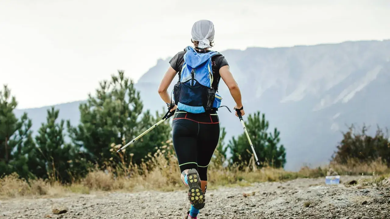 Frauen-Speed-Hiking mit Rucksack und Stöcken auf einem Bergpfad, umgeben von Bäumen und mit Blick auf Berge.