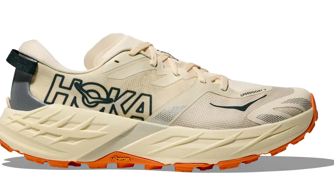 Beiger Hoka Speedgoat 7 Trailrunningschuh mit orangefarbener Vibram-Sohle und dunklem Hoka-Logo.