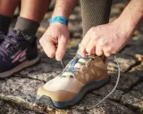 Hände binden Schnürsenkel eines beigefarbenen Schuhs, bereit für den Trail. Daneben ein lila Schuh, der verschiedene wanderschuhe kategorien zeigt.