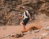 Läufer beim Marathon des Sables wirbelt Sand auf. Ein Mann mit Rucksack und Stirnlampe kämpft sich durch die Wüste.