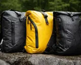 Drei wasserdichte Rucksäcke mit PU-Beschichtung stehen im Regen auf Felsen. Einer ist gelb, die anderen beiden schwarz.