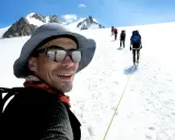 Mann mit Sonnenbrille und Hut, lachend, während er mit einer Gruppe auf einem schneebedeckten Berg wandert. Trail training in den Alpen.