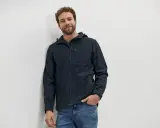 Mann mit lockigem Haar trägt eine dunkelblaue Softshelljacke und Jeans. Die Jacke ist bereit für die Wäsche.