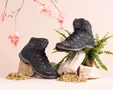 Zwei schwarze Wanderschuhe von LOWA, auf Steinen und Kies arrangiert, mit zarten rosa Blüten und Farn im Hintergrund.