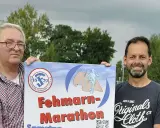 Zwei Männer präsentieren stolz ein Plakat für den Fehmarn Marathon. Das Logo des Sportvereins Fehmarn ist zu sehen.