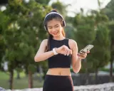 Lächelnde junge Frau mit Kopfhörern und Smartphone, die ihre Smartwatch prüft. Sie trägt atmungsaktive Kleidung für ihr Training im Freien.