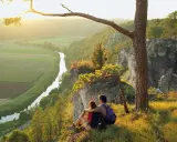 Ein Paar genießt den Sonnenuntergang nach besonderen Wanderungen auf einem Felsvorsprung mit Blick auf ein Tal mit Fluss und Feldern.