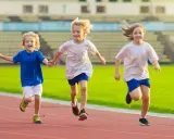 Drei lachende Kinder beim Lauftraining auf einer roten Laufbahn. Sie rennen nebeneinander, die Arme ausgestreckt.