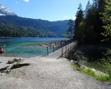 Ein Steg führt über das klare Wasser des Eibsees, ideal für den Eibsee Rundweg. Im Hintergrund Berge und ein rotes Boot.