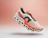 Ein weißer Laufschuh mit orangefarbenen Akzenten, der Cloudmonster 2 Herren, schwebt vor einem sanften Farbverlauf.