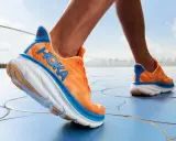Ein Läufer testet die Hoka Clifton 9 Schuhe in leuchtendem Orange mit blauen Akzenten, die auf einem blauen Boden unterwegs sind.