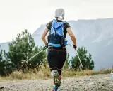 Frauen-Speed-Hiking mit Rucksack und Stöcken auf einem Bergpfad, umgeben von Bäumen und mit Blick auf Berge.