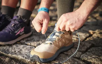 Hände binden Schnürsenkel eines beigefarbenen Schuhs, bereit für den Trail. Daneben ein lila Schuh, der verschiedene wanderschuhe kategorien zeigt.