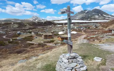 Wanderwegweiser im Rondane Nationalpark, mit grasbewachsenen Hütten und schneebedeckten Bergen im Hintergrund.