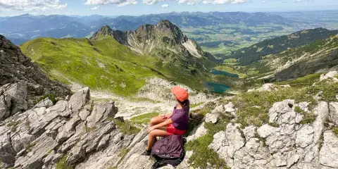Ein Mädchen mit Rucksack genießt die Aussicht auf die Berge bei Oberstdorf. Ein leichtes Wandern mit toller Belohnung.