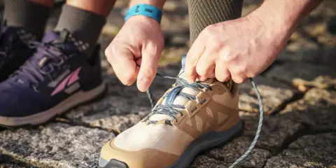 Hände binden Schnürsenkel eines beigefarbenen Schuhs, bereit für den Trail. Daneben ein lila Schuh, der verschiedene wanderschuhe kategorien zeigt.