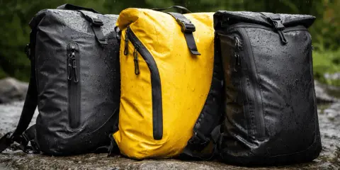 Drei wasserdichte Rucksäcke mit PU-Beschichtung stehen im Regen auf Felsen. Einer ist gelb, die anderen beiden schwarz.