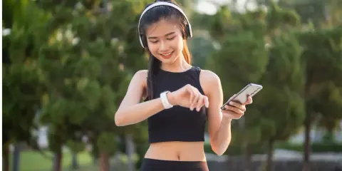 Lächelnde junge Frau mit Kopfhörern und Smartphone, die ihre Smartwatch prüft. Sie trägt atmungsaktive Kleidung für ihr Training im Freien.
