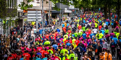 Große Menschenmenge beim Firmenlauf Essen, viele Läufer in bunten Trikots auf einer Straße, umgeben von Bäumen und Gebäuden.
