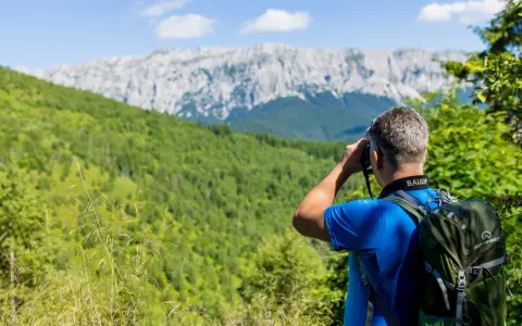 Mann mit Rucksack beim Karpaten Wandern, blickt durch Fernglas auf bewaldete Hänge und felsige Gipfel.