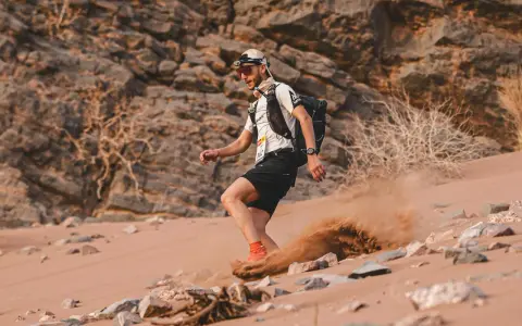 Läufer beim Marathon des Sables wirbelt Sand auf. Ein Mann mit Rucksack und Stirnlampe kämpft sich durch die Wüste.