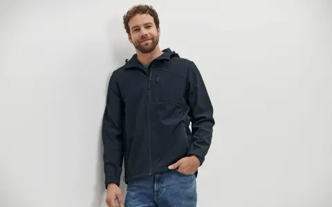 Mann mit lockigem Haar trägt eine dunkelblaue Softshelljacke und Jeans. Die Jacke ist bereit für die Wäsche.