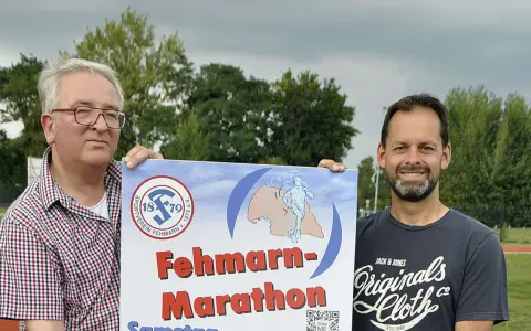 Zwei Männer präsentieren stolz ein Plakat für den Fehmarn Marathon. Das Logo des Sportvereins Fehmarn ist zu sehen.
