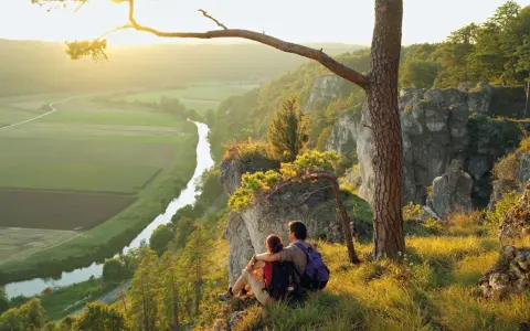Ein Paar genießt den Sonnenuntergang nach besonderen Wanderungen auf einem Felsvorsprung mit Blick auf ein Tal mit Fluss und Feldern.