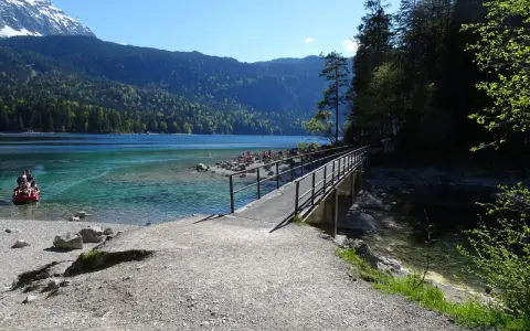 Ein Steg führt über das klare Wasser des Eibsees, ideal für den Eibsee Rundweg. Im Hintergrund Berge und ein rotes Boot.