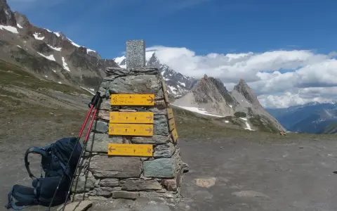 Wegweiser für die Tour du Mont Blanc Etappen: Rucksack und Stöcke liegen bereit. Bergpanorama mit blauen Himmel und Wolken.
