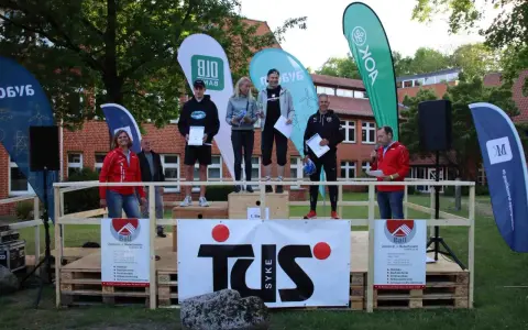 Siegerehrung beim Hachelauf Syke: Läufer auf dem Podium, Moderation und Sponsoren-Banner im Hintergrund.