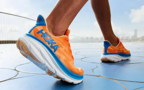 Ein Läufer testet die Hoka Clifton 9 Schuhe in leuchtendem Orange mit blauen Akzenten, die auf einem blauen Boden unterwegs sind.