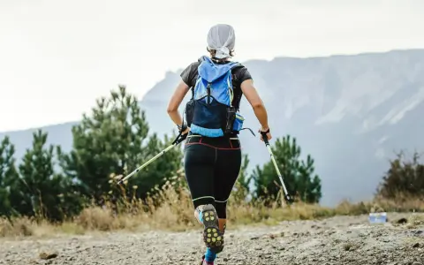 Frauen-Speed-Hiking mit Rucksack und Stöcken auf einem Bergpfad, umgeben von Bäumen und mit Blick auf Berge.